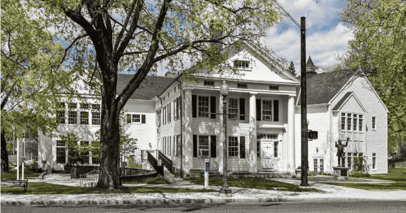 A historic white wooden building with columns, surrounded by trees in a small town setting.