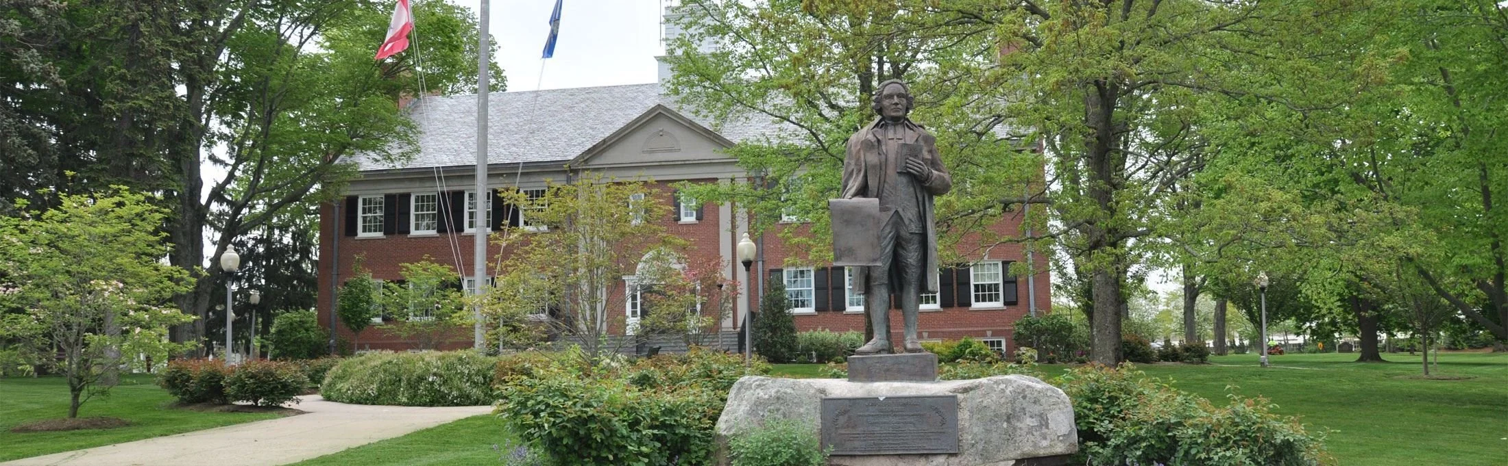 Statue of a historical figure holding a book, situated in a green park with trees and a brick building in the background.