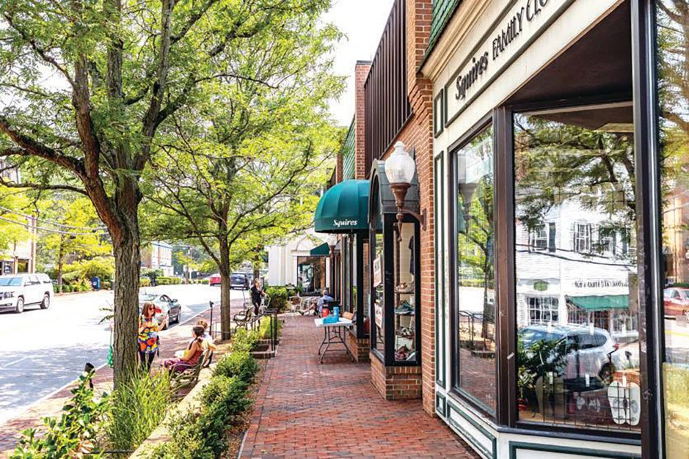 Downtown sidewalk with storefronts, including 'Squires Family' shop, brick buildings, trees, and pedestrians.