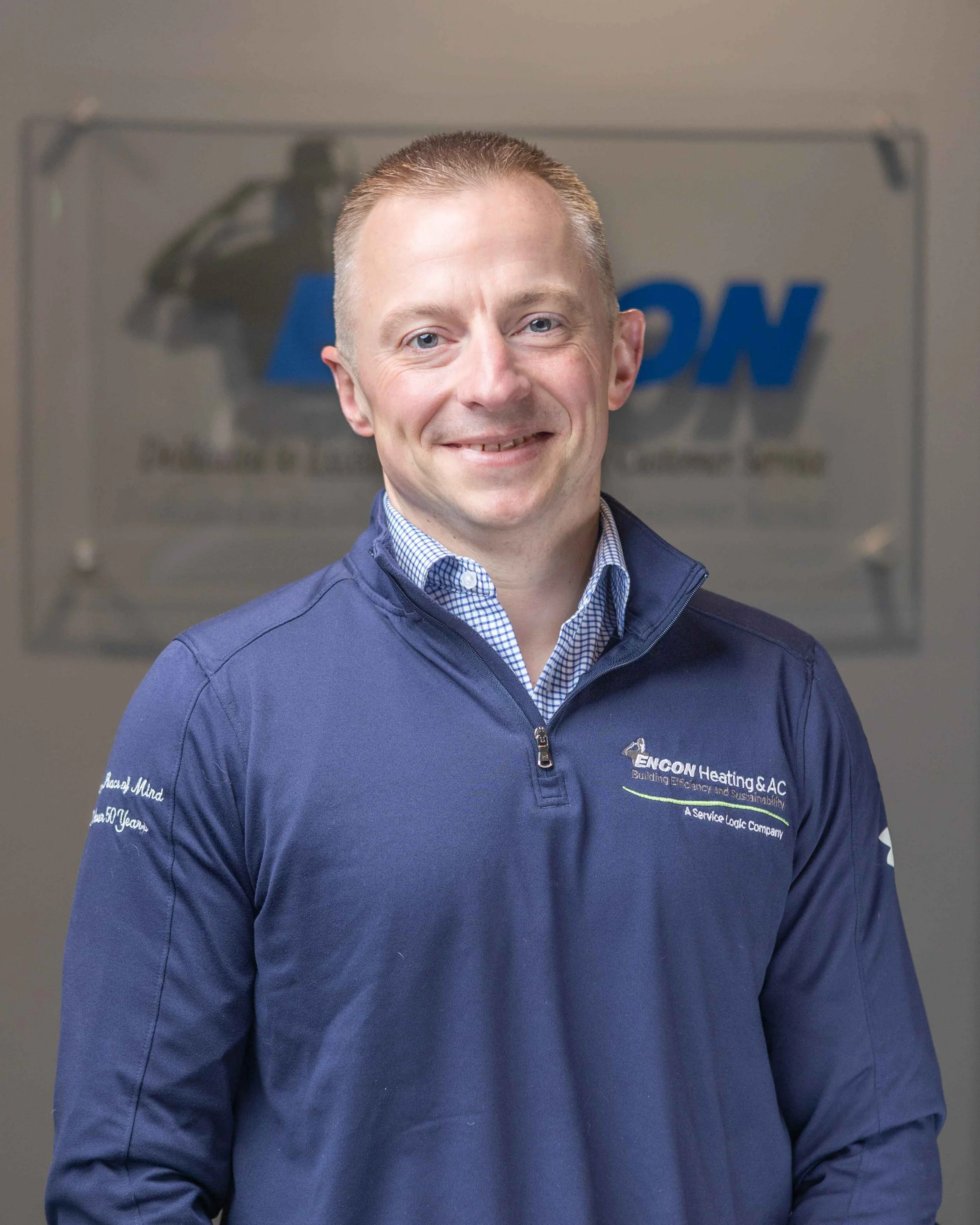 A man in a blue jacket with company logo, smiling indoors with a blurred company sign in background.