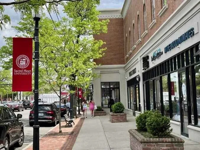 A sidewalk scene with trees and parked cars along a street in front of storefronts including Sacred Heart University. Two women in pink dresses walk down the sidewalk.