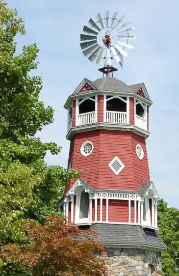 A red and white old-fashioned water tower with a windmill on top, set against a blue sky and surrounded by green trees.