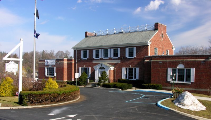 A brick building with white-framed windows and a greenish roof, surrounded by bushes and a small snow pile, with a parking lot and flagpoles in front, on a partly cloudy day.