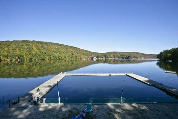 A calm lake with a floating dock and a few people sitting on the shore, surrounded by green hills under a clear blue sky.