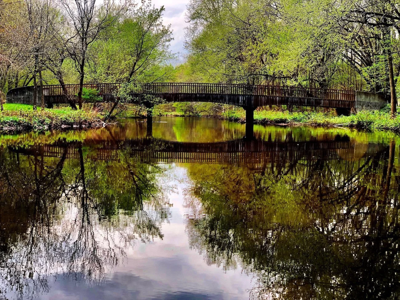 A wooden bridge over a calm river in a park with green trees and reflections in the water.
