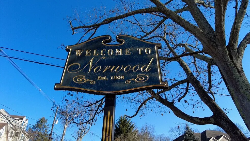 A welcome sign for Norwood, established in 1905, hanging from a tree with bare branches, against a clear blue sky.