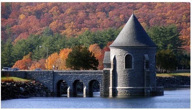A stone castle-like structure with a conical roof by a body of water, surrounded by colorful fall foliage.