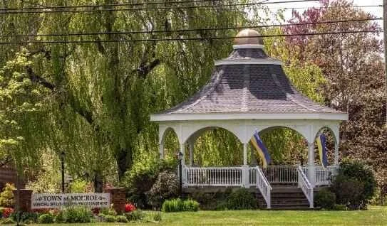 White gazebo with a dark roof in a park, surrounded by green trees and a lawn, with a small sign and flowers nearby.