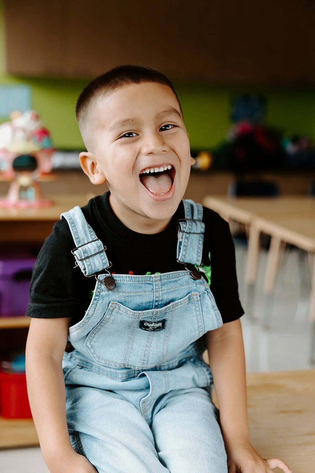 Un garçon souriant assis sur une surface en bois dans une salle de classe, portant un t-shirt noir et des dungarees en denim clair, dans un style de photographie scolaire au Québec.