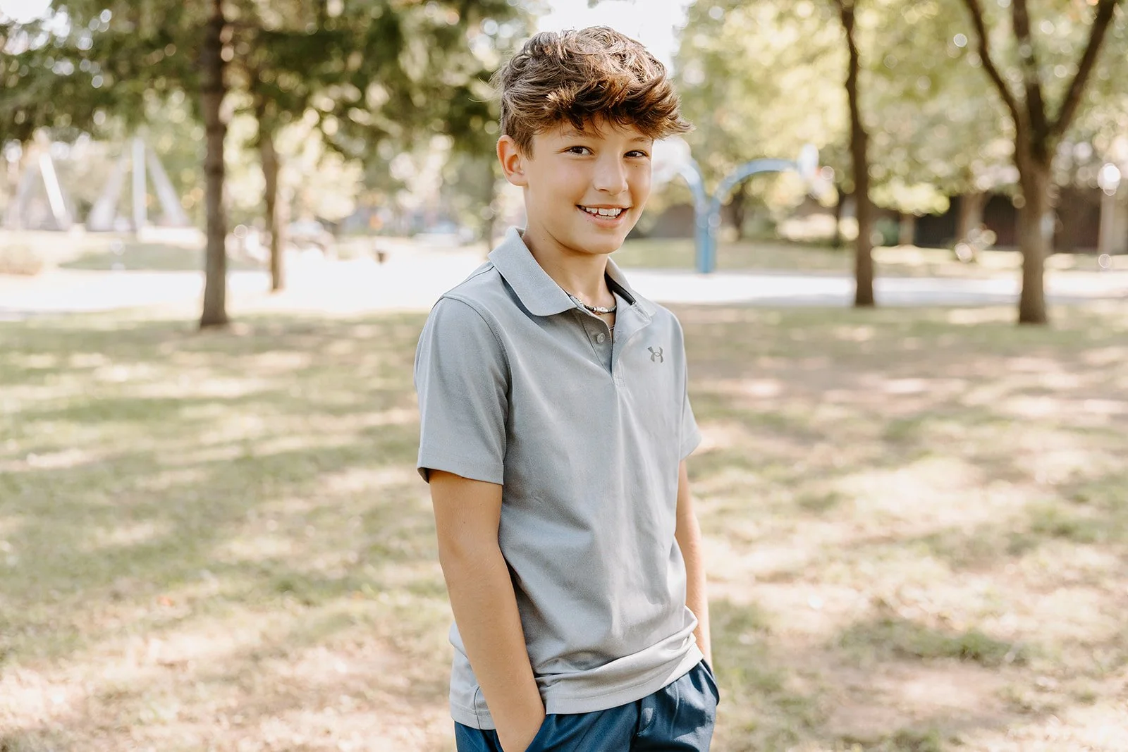 Un garçon souriant en polo gris se tient dans un parc en lumière naturelle, dans un style de photographie scolaire réalisée par des photographes au Québec.