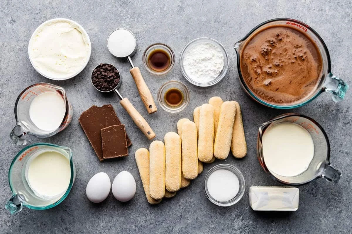 Ingredients for chocolate ladyfingers on a gray surface, including eggs, milk, chocolate, sugar, flour, cocoa, cream, and a chocolate mixture.