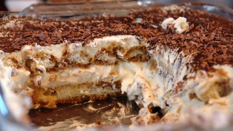 A layered homemade tiramisu dessert in a glass dish with visible cream, sponge cake, and chocolate shavings on top.