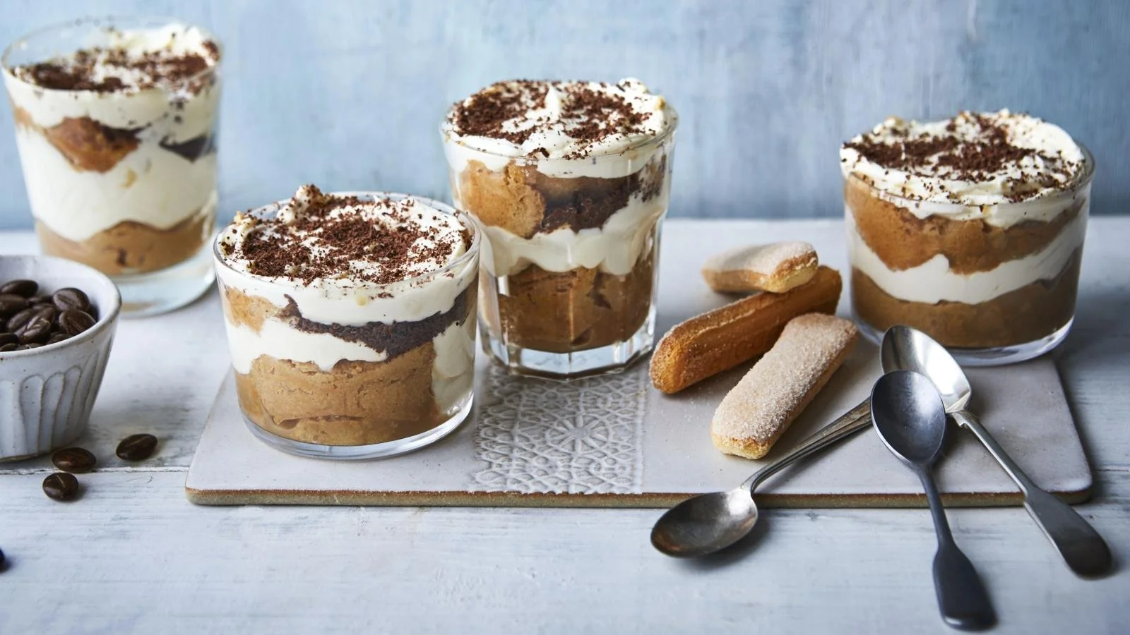 Four glasses of tiramisu with cream, coffee-soaked cake, and chocolate sprinkles on top, placed on a white board with cookies and spoons nearby, on a white wooden surface.
