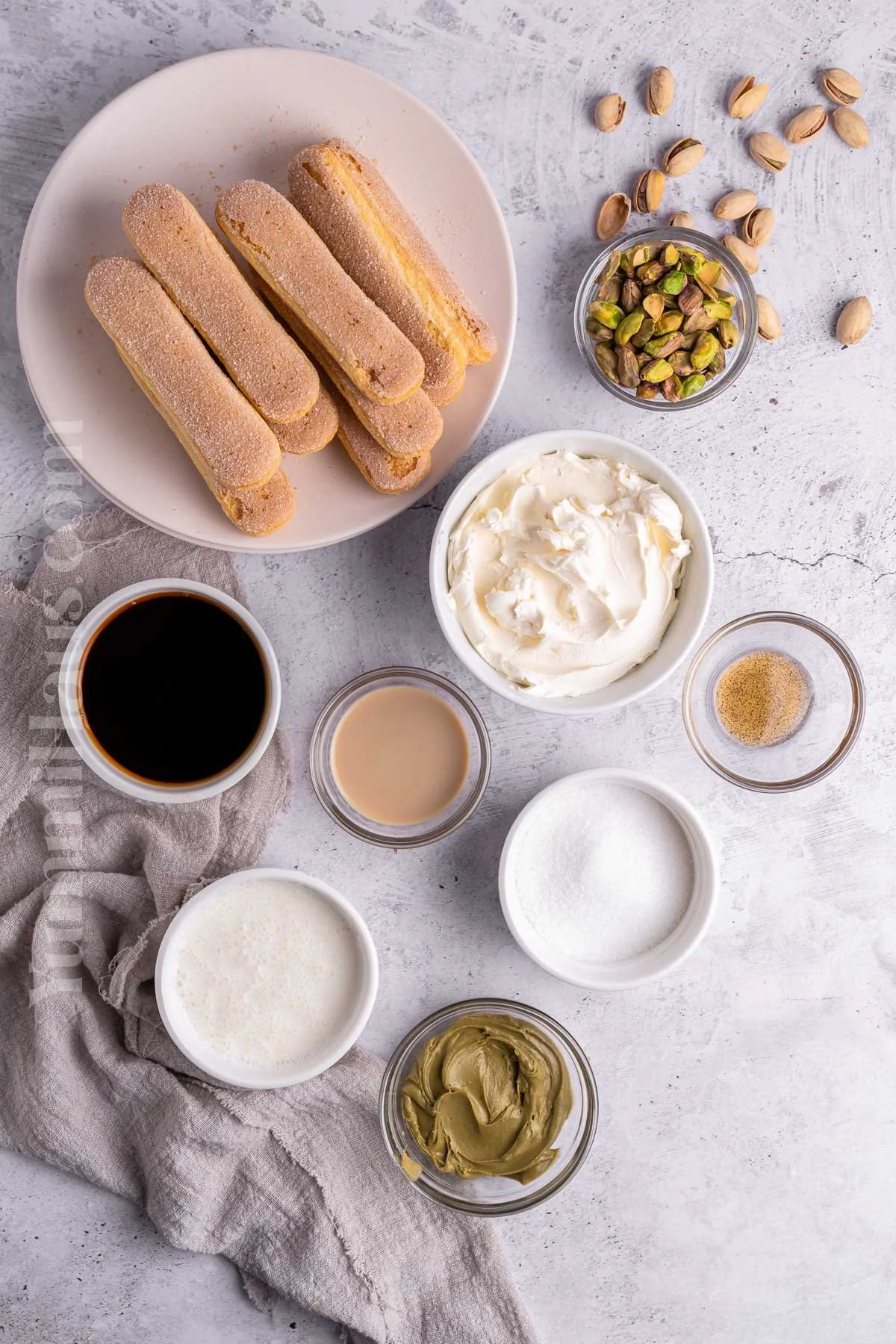 Assorted ingredients for making tiramisu including ladyfinger cookies, chopped pistachios, mascarpone cheese, coffee, sweetened condensed milk, sugar, heavy cream, and green tea paste, arranged on a white surface.