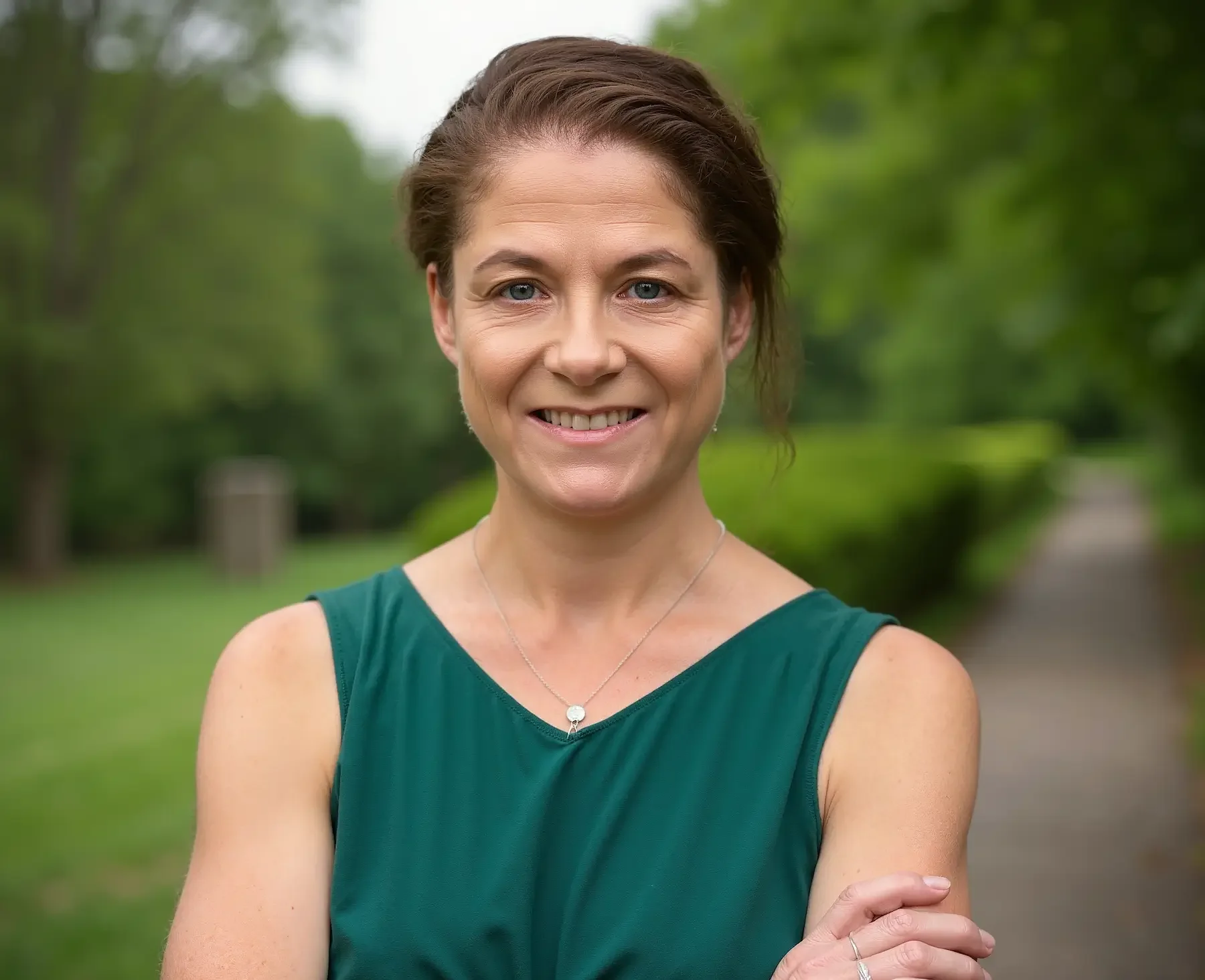 A woman standing outdoors on a path in a park, smiling at the camera, with trees and greenery in the background.