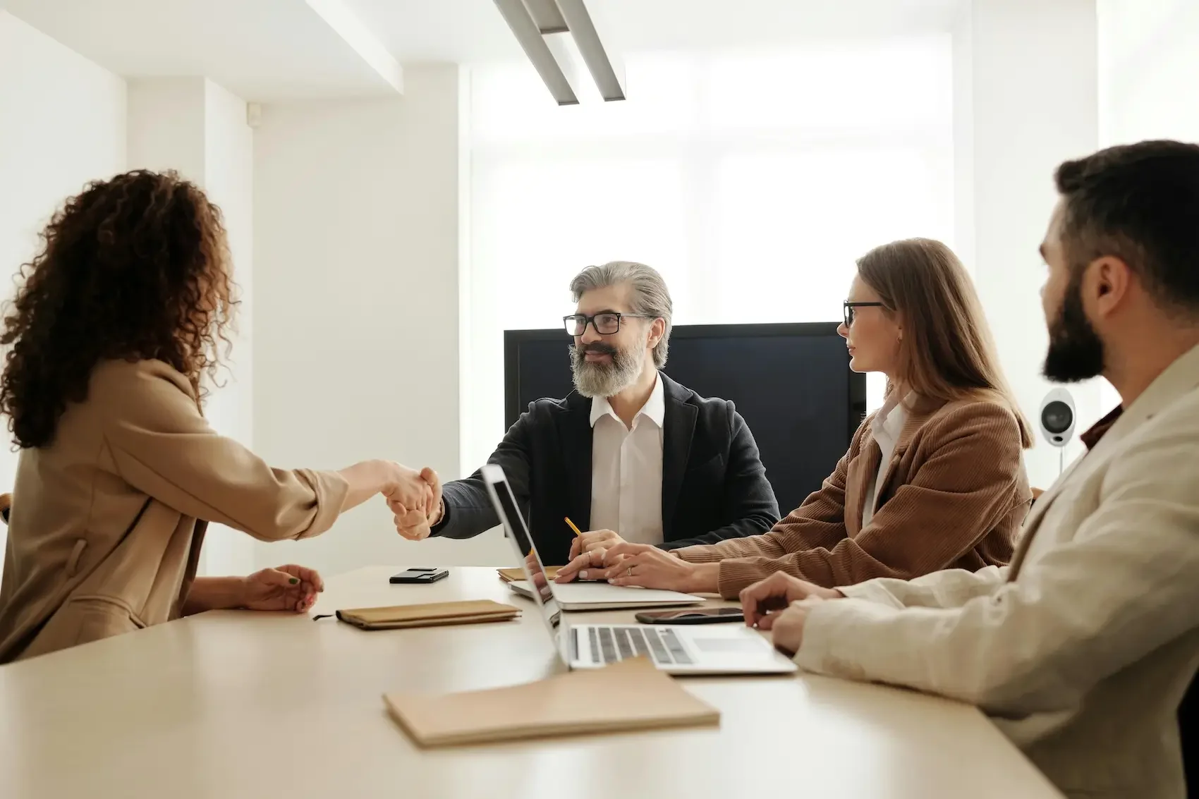 Business meeting with four people, two men and two women, engaging in handshake and discussion in a modern office.