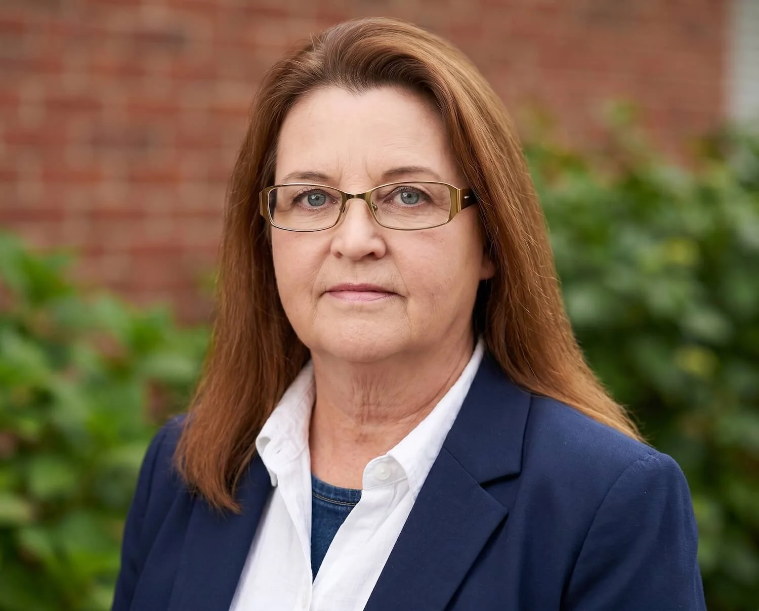A woman with glasses stood outside with a wall and greenery behind her