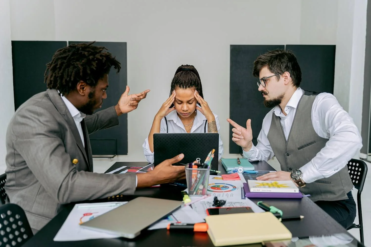 Three people in office attire sit at a table. Two are arguing and gesticulating, the other has their head in their hands