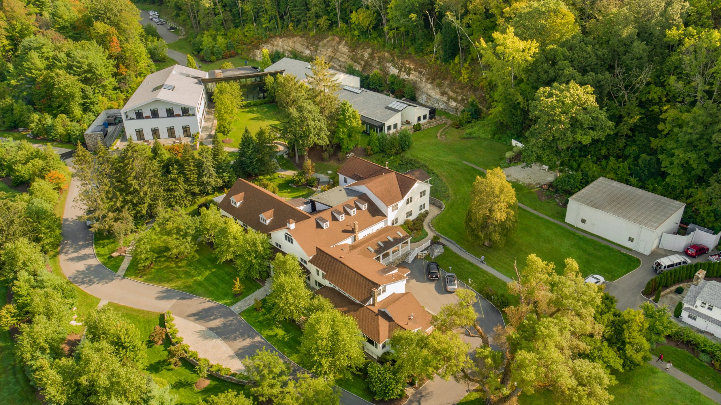 An aerial view of a residential area with large houses, trees, green lawns, and a winding road surrounded by wooded hills.