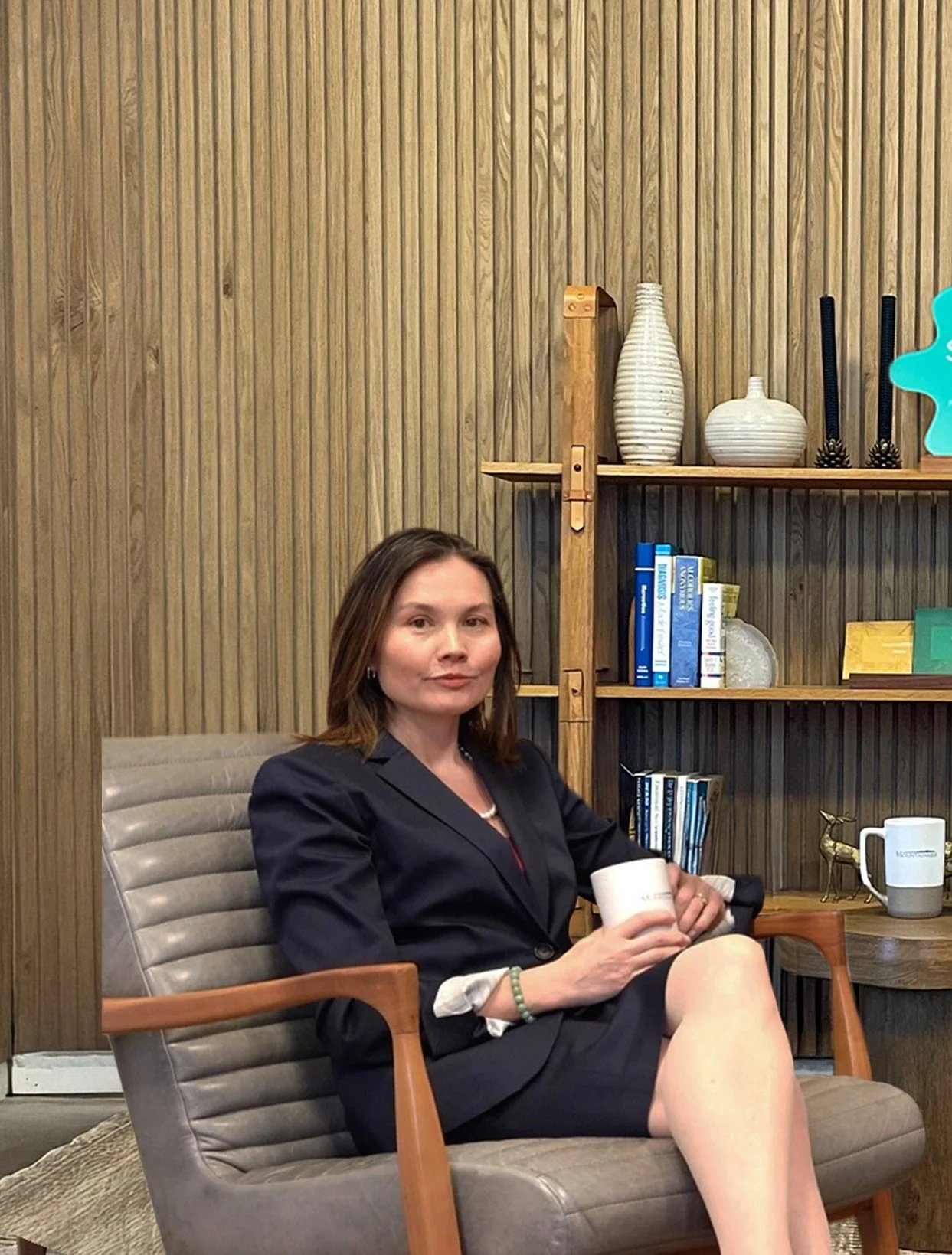 A woman in a black blazer sitting on a gray armchair, holding a coffee mug, in a room with wood panel walls, bookshelf, and decorative items.