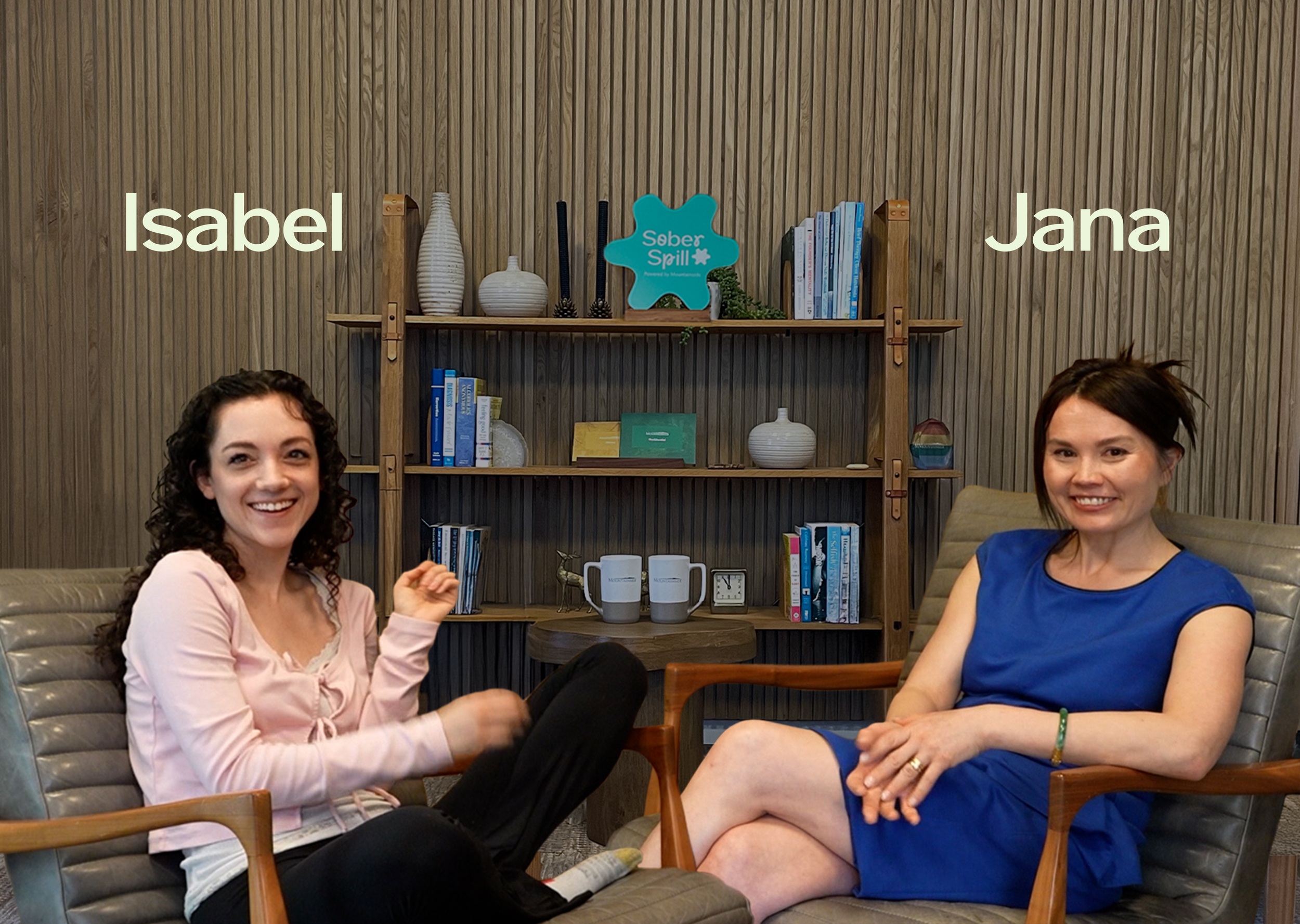 Two women sitting in chairs in a room with a wooden bookshelf in the background. The woman on the left has curly hair, wearing a pink top and black pants. The woman on the right has straight hair, wearing a blue dress. The room has books, decorative items, and a sign that says 'Seber Spill' on the bookshelf.