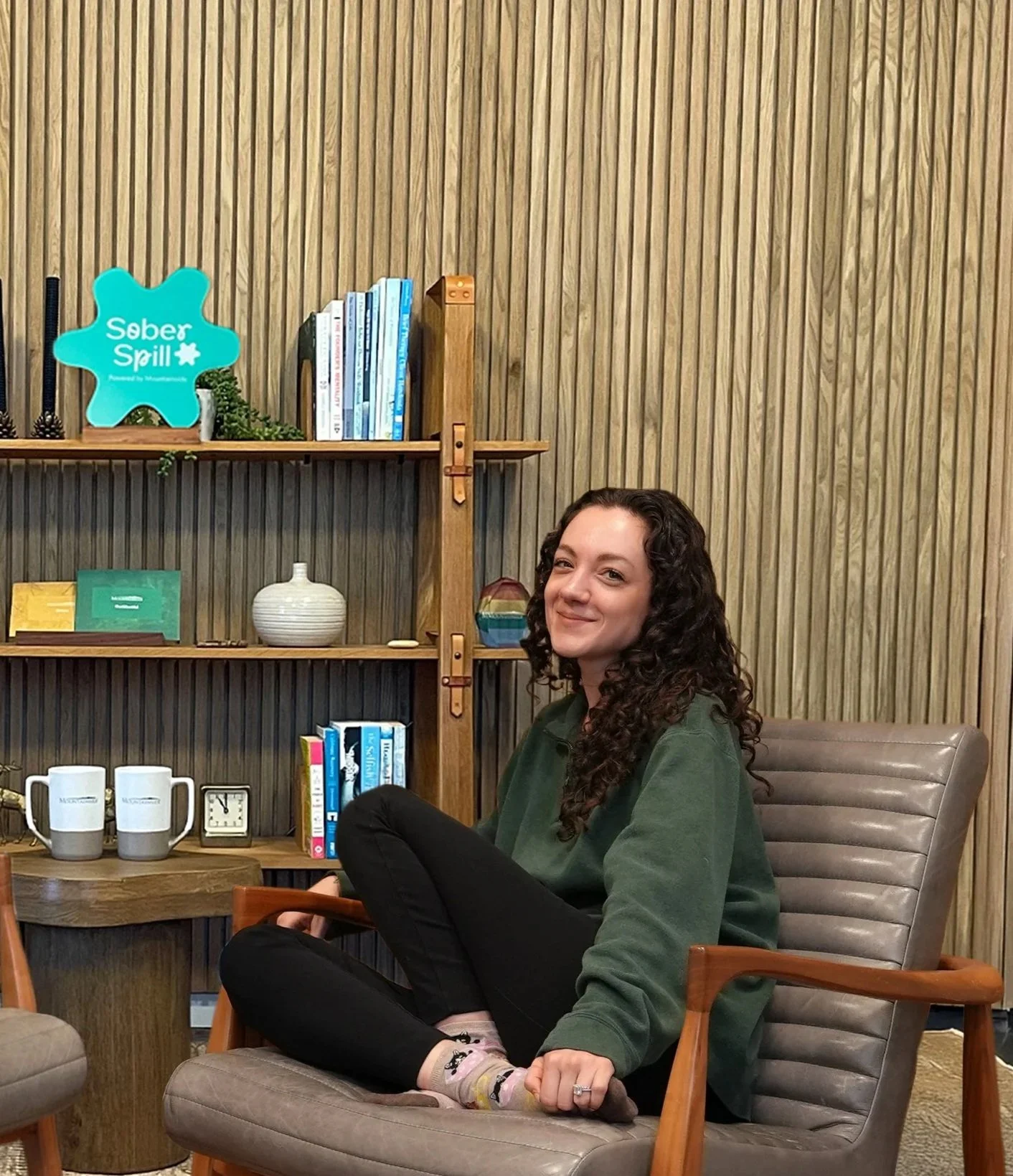 A woman with curly dark hair smiling while sitting in a lounge chair in a cozy room with wooden panel walls, a bookshelf, and decorative items.