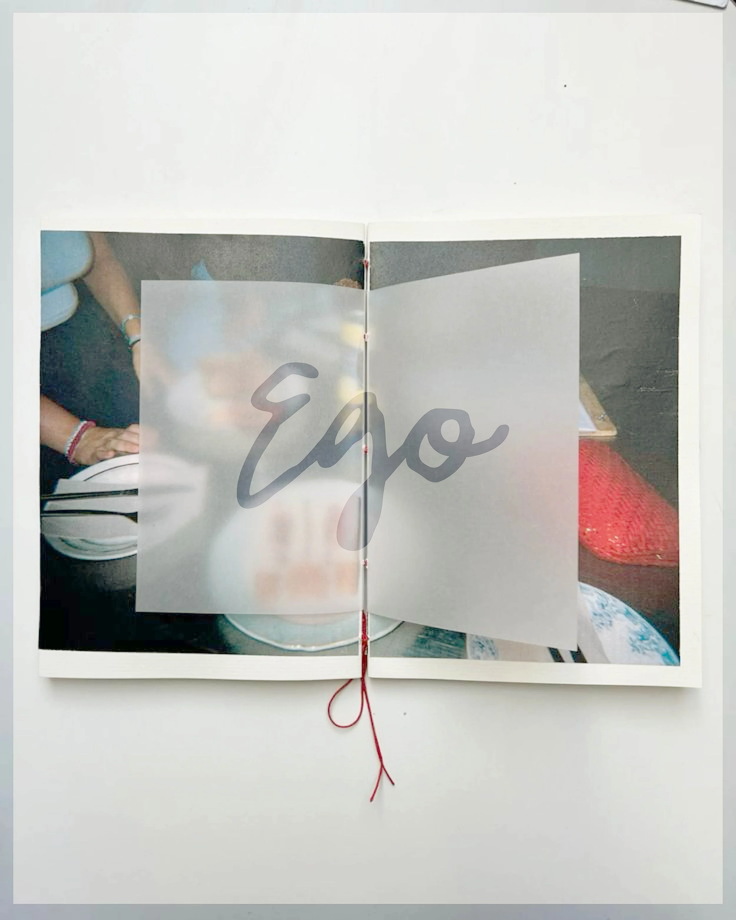 Open book with a photograph of a table setting showing someone and a red purse, partially obscured by a semi-transparent page with the word 'Eyo' written across it.