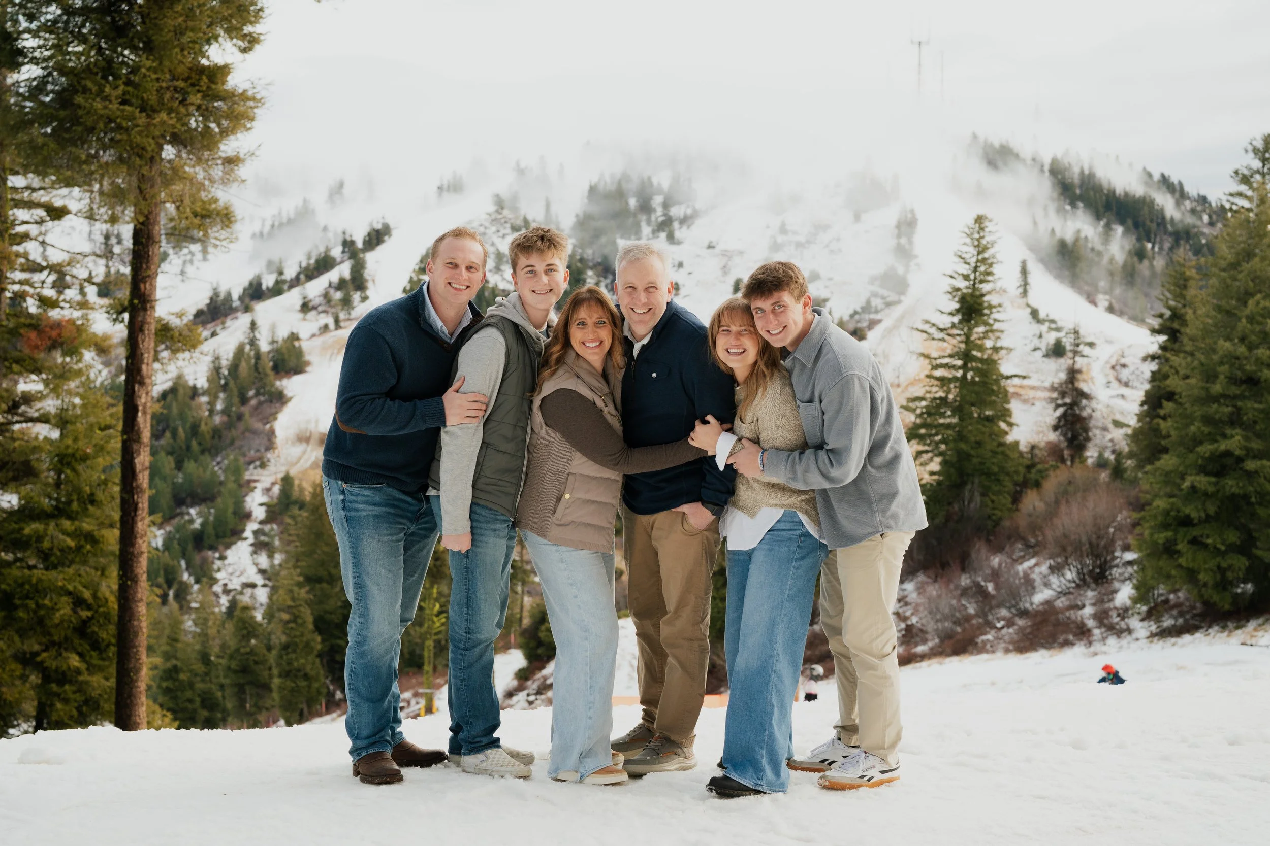 A group of six people, five adults and one child, standing on snow in a mountain landscape with pine trees and snow-covered slopes, smiling and embracing each other.