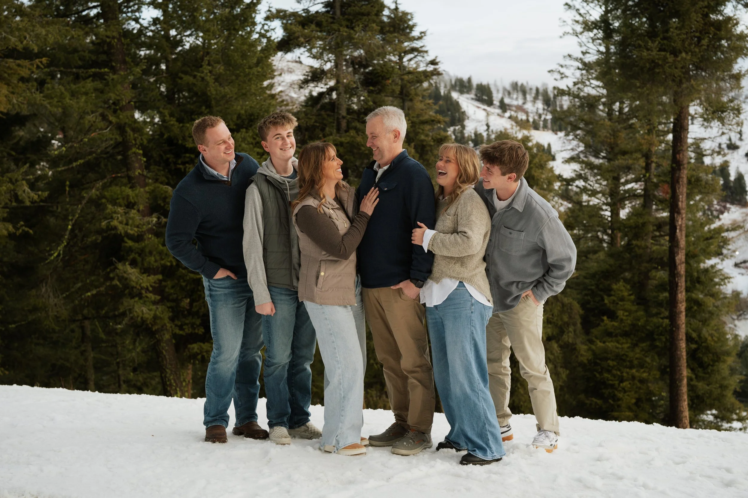Group of seven people standing on snow-covered ground outdoors, smiling and interacting joyfully, with pine trees and snow-covered hills in the background.