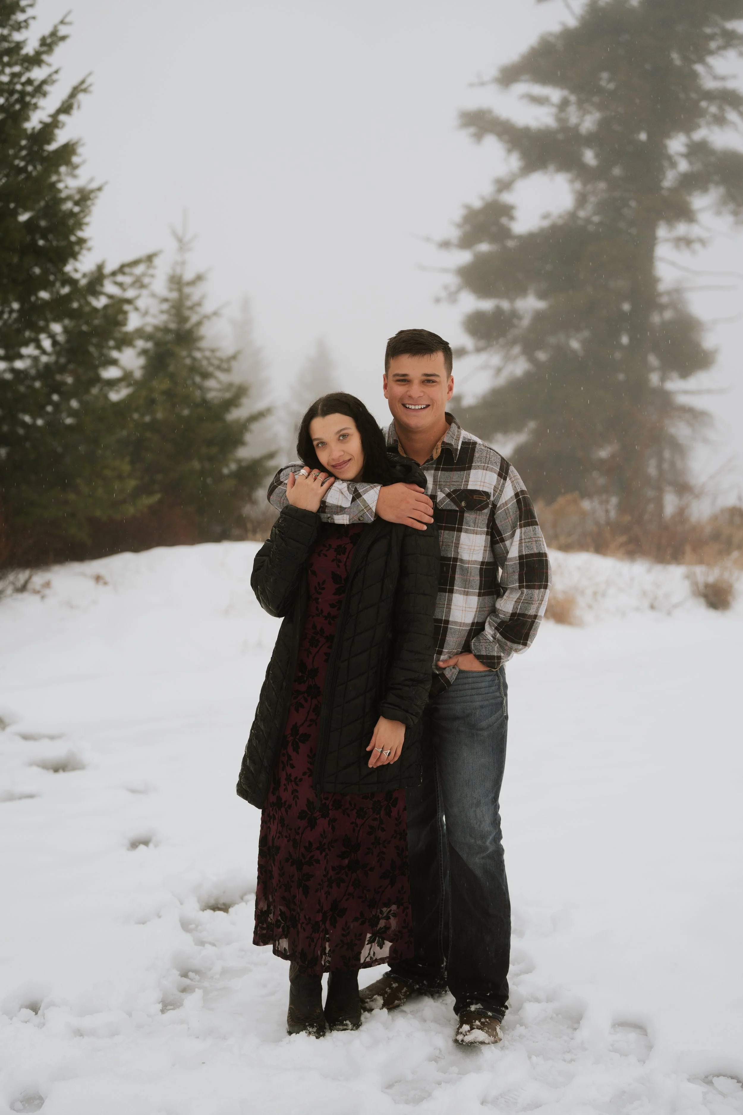 A smiling couple standing close together outdoors in a snowy landscape with trees in the background. The man has his arm around the woman's shoulders, and she is leaning her head on his chest.