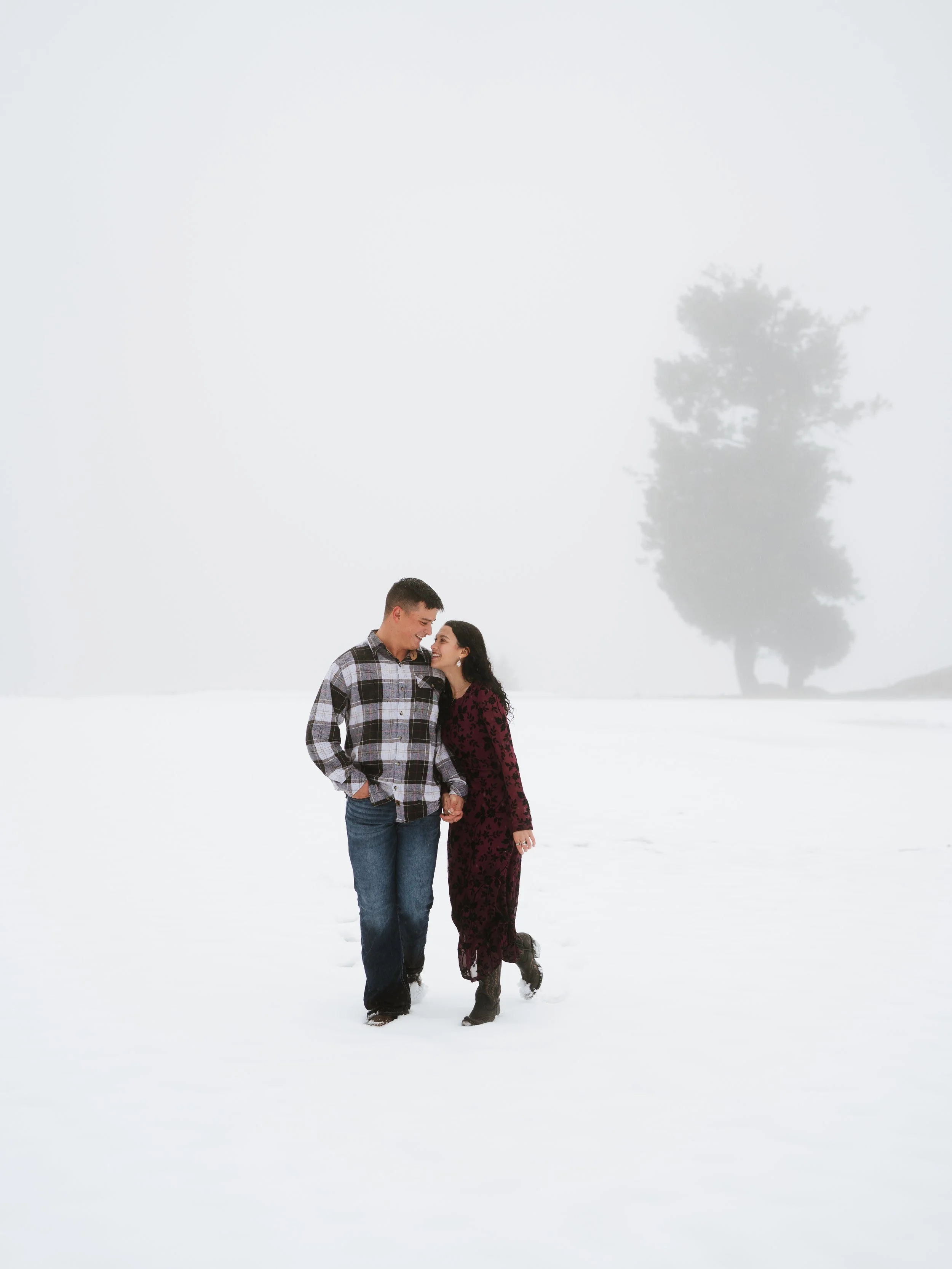 A couple holding hands and smiling in a snowy outdoor setting with a foggy background and a large tree in the distance.