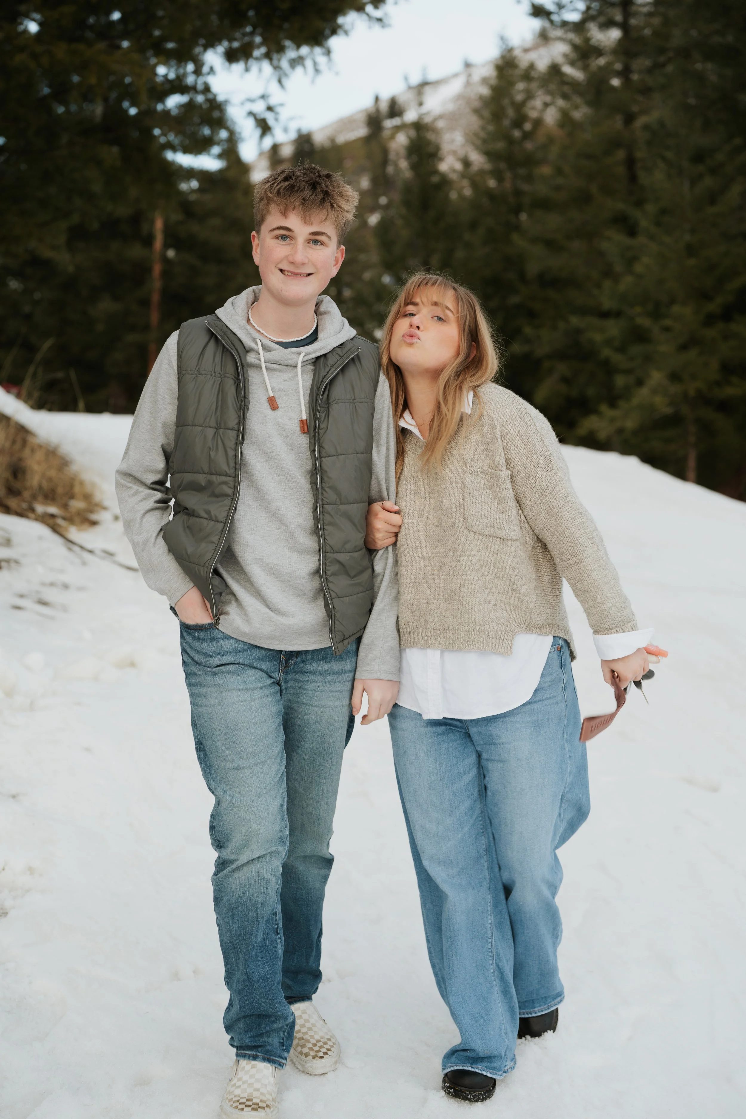 A young man and woman walking in the snow outdoors, surrounded by evergreen trees and mountains in the background. The man is smiling with one hand in his pocket, wearing a gray hoodie, a green vest, and jeans. The woman is making a kissy face with her lips puckered, holding a small brown item, and is dressed in a beige sweater, white shirt, and wide-leg jeans.