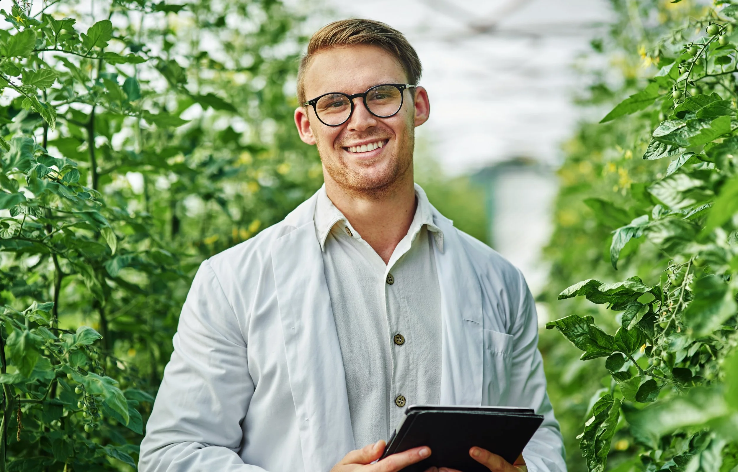 A smiling man in glasses and a white lab coat holds a clipboard in a greenhouse filled with green plants.