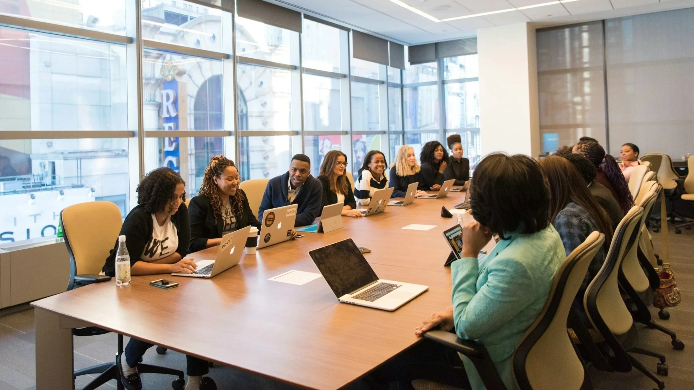 A diverse group of people sitting around a conference table in a modern office, working on laptops and tablets, with windows showing an urban cityscape in the background.