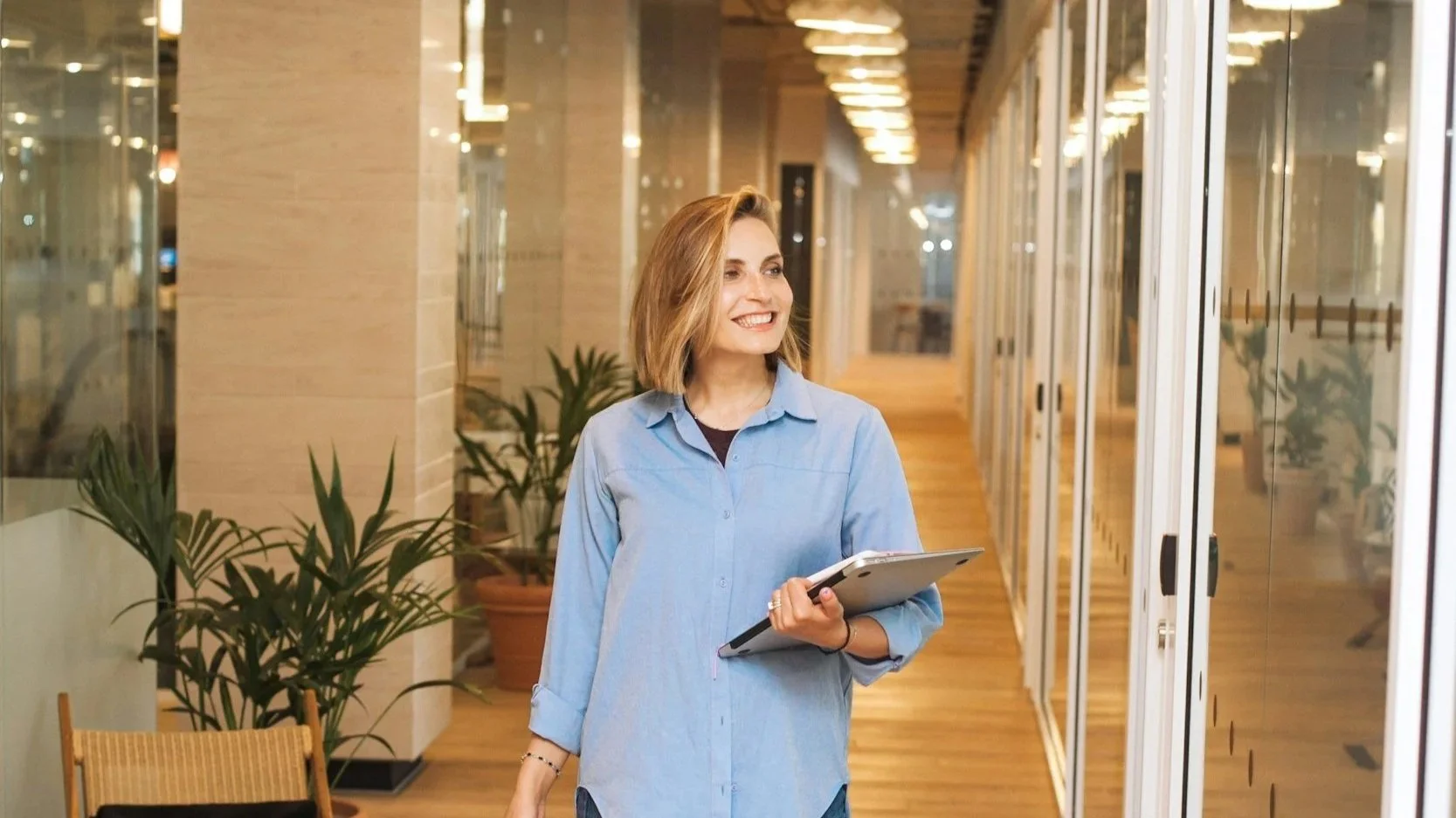 A woman with shoulder-length blonde hair, wearing a light blue button-up shirt, holding a tablet, walking in a modern office corridor with glass walls and potted plants.