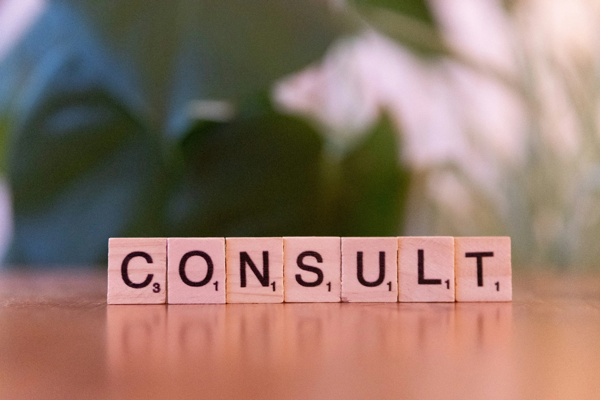 Scrabble tiles spell out the word 'CONSULT' on a wooden surface, with leaves and blurry flowers in the background.