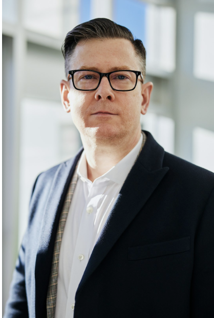 Portrait of a man wearing glasses and a dark blazer, standing indoors near large windows.