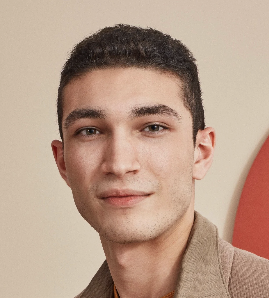 A young man with short dark hair and light skin, wearing a tan jacket, smiling gently against a beige background.
