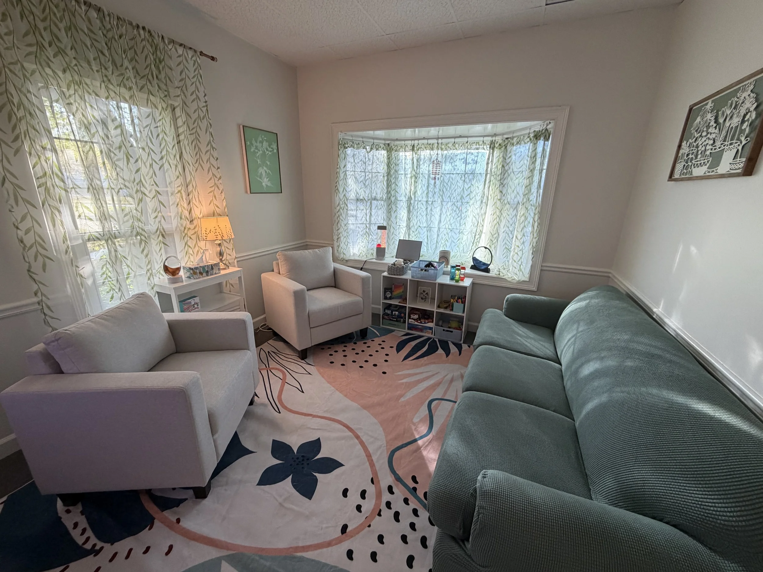Therapy room for children and teens with ocd, anxiety, and other disorders in SNellville. The room is cozy with a green couch on one side and two white chairs on the other. there is a pink and white floral rug on teh ground, and two large windows.