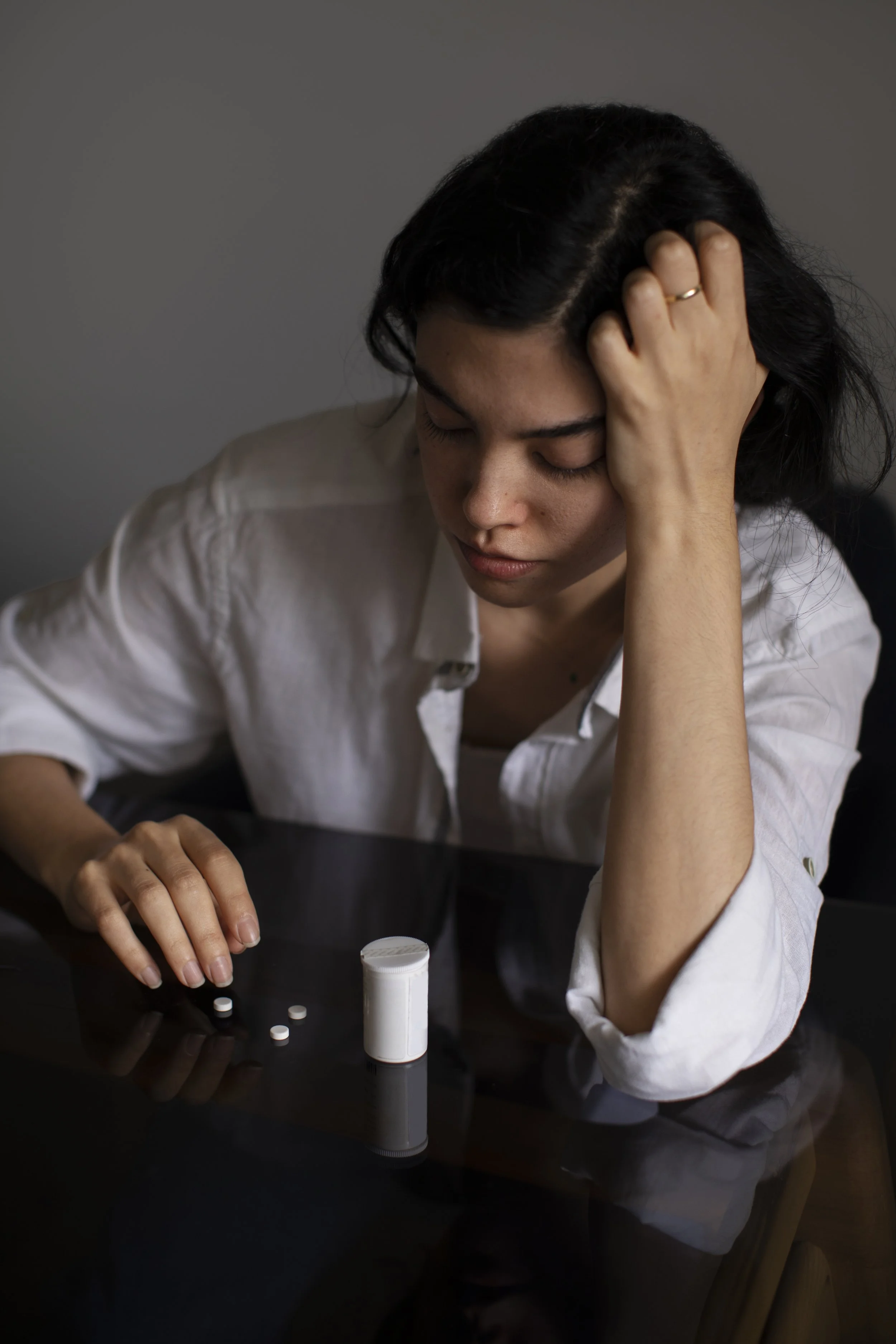 A woman with dark hair and a white shirt sitting at a dark table, holding pills in her hand and looking down with a distressed expression, with a bottle of pills on the table. Credits for FreePik