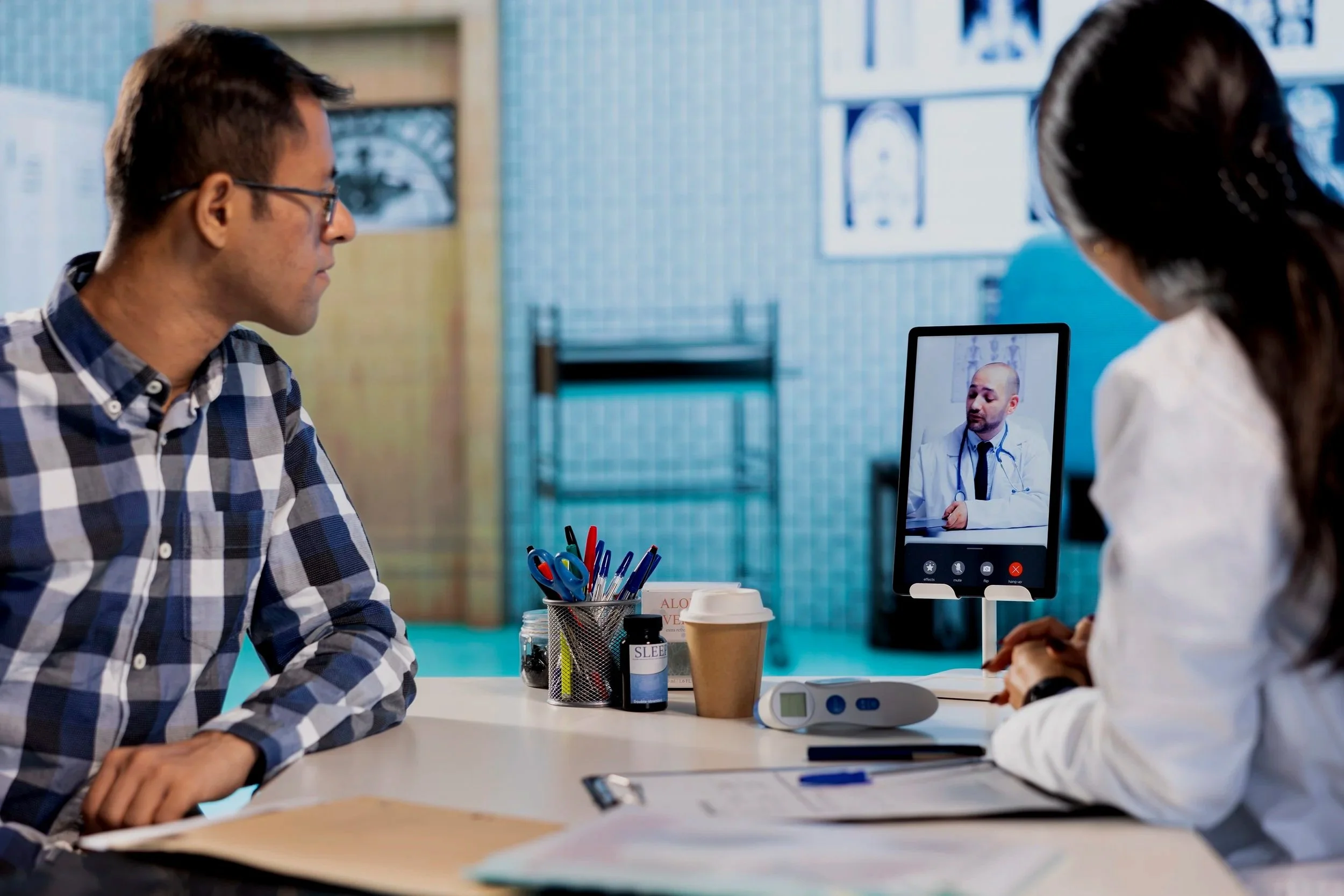 A man and a woman sitting at a desk, having a video call with a doctor on a tablet. The man is wearing a plaid shirt, and the woman is in a white coat. The desk has medical supplies and a coffee cup. Credits for FreePik