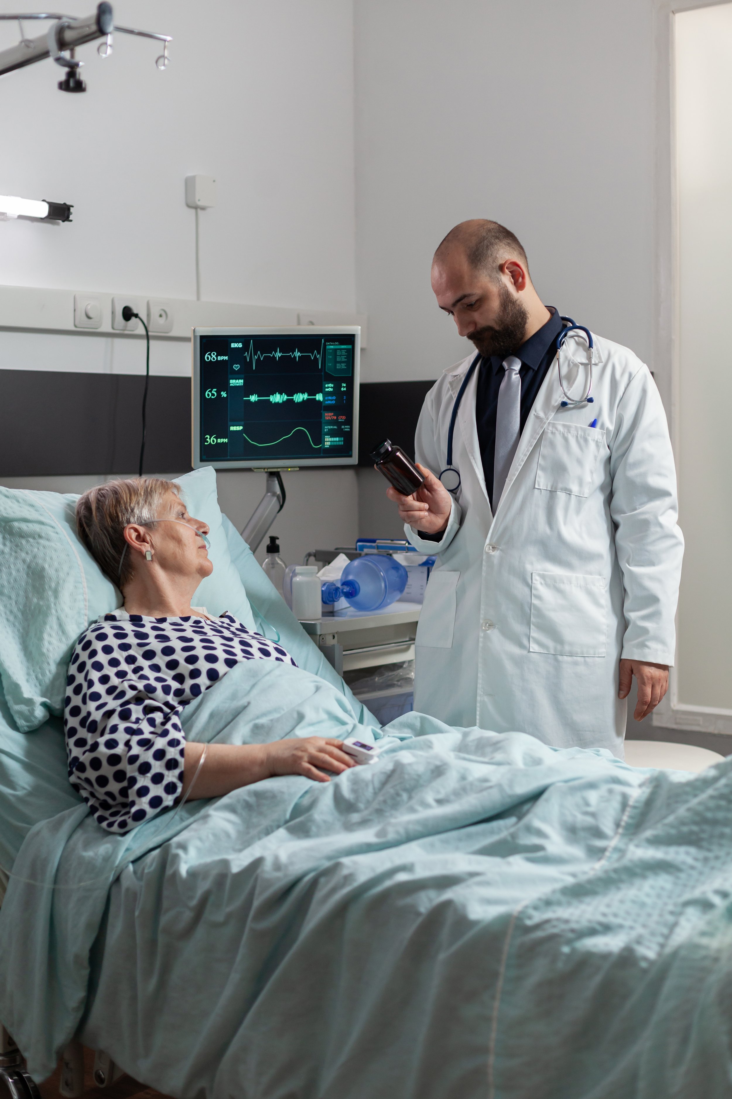 A doctor standing next to a patient in a hospital bed, reviewing medication or notes. The patient, an elderly woman, is lying in bed and connected to medical devices, with a monitor displaying vital signs behind her. Credits for FreePik