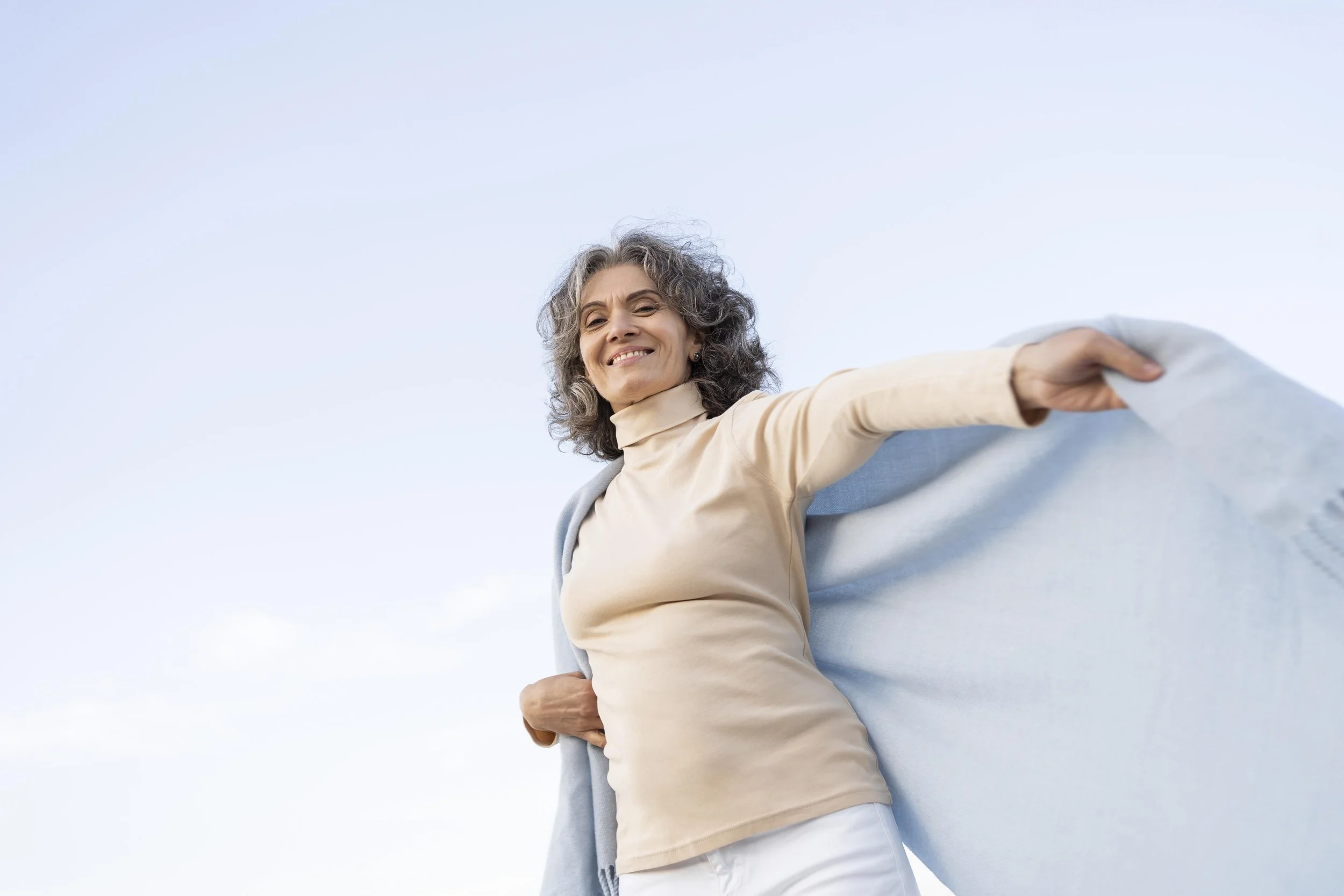 A woman with curly gray hair smiling outdoors, holding her light blue jacket open as if flying it like a cape against a clear sky. Credits for FreePik