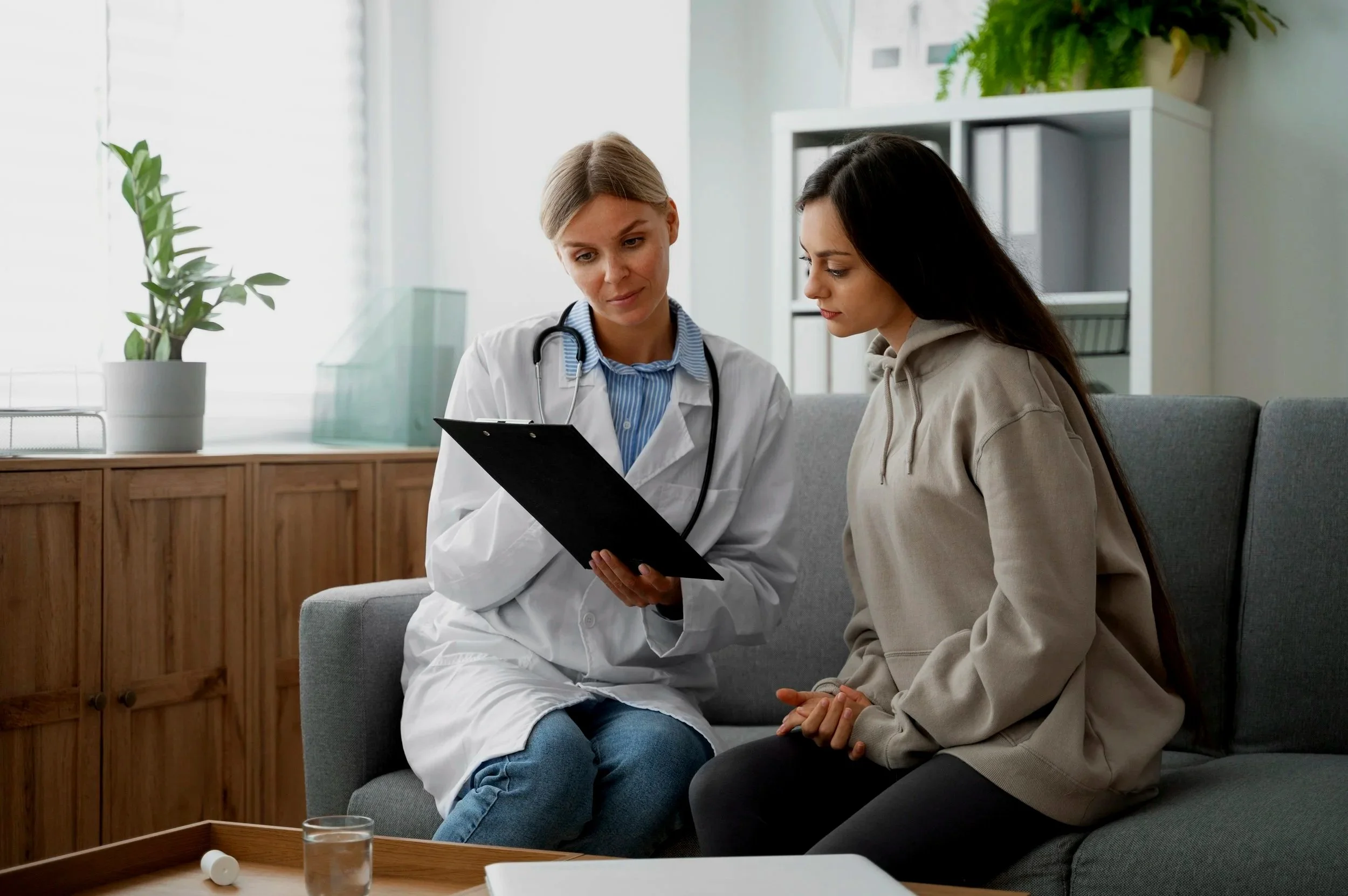 Medical professional in white coat and stethoscope talking with young woman in beige hoodie in a clinic or mental health office. Credits for FreePik