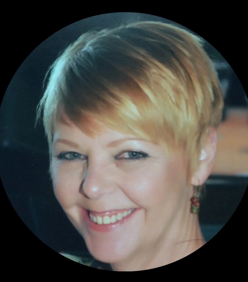 Close-up of a smiling woman with short blonde hair, blue eyes, and earrings, inside a circular frame.