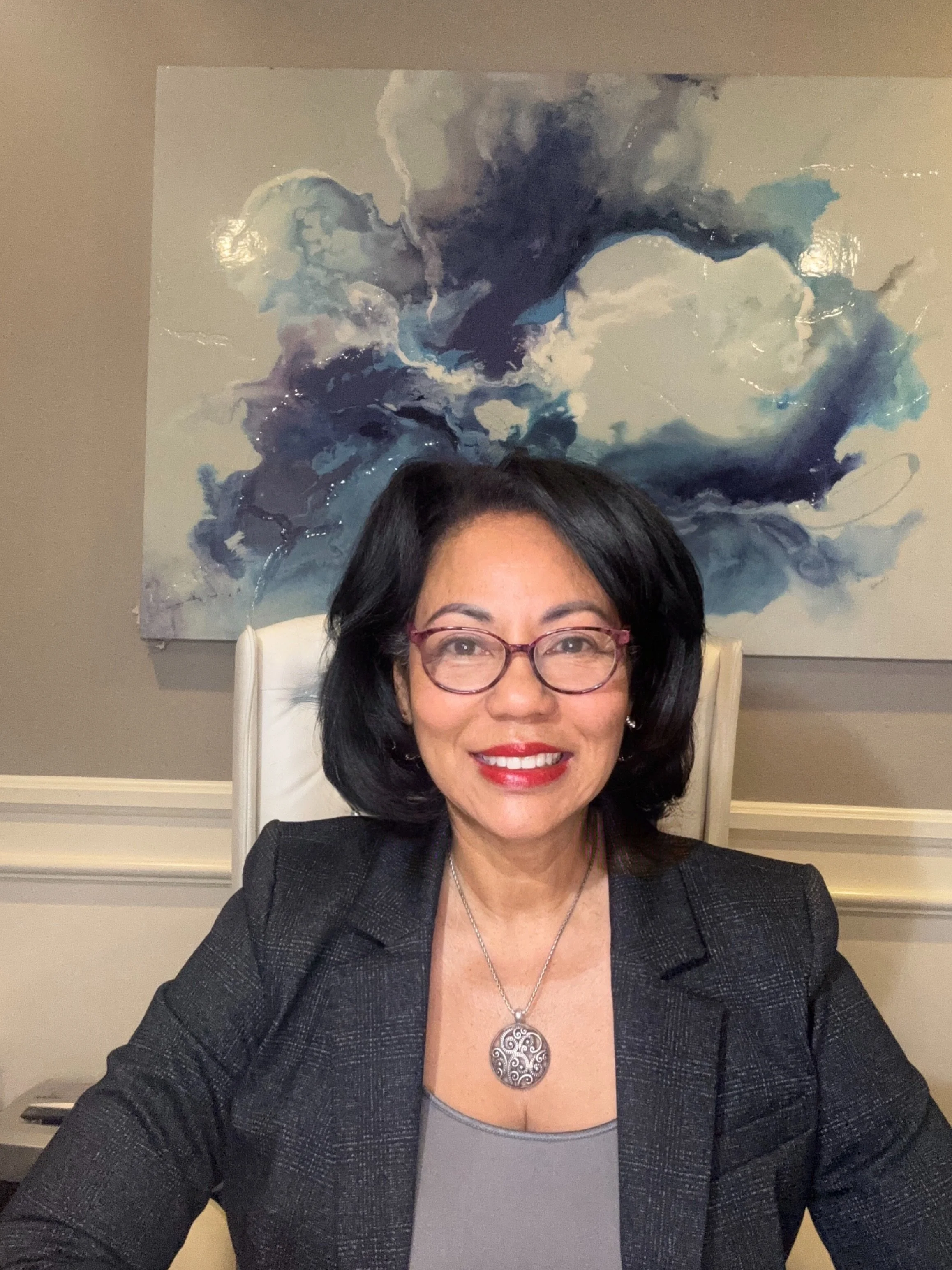 A woman with black hair, glasses, and red lipstick smiling, sitting at a desk with a painting of abstract blue and white clouds behind her.