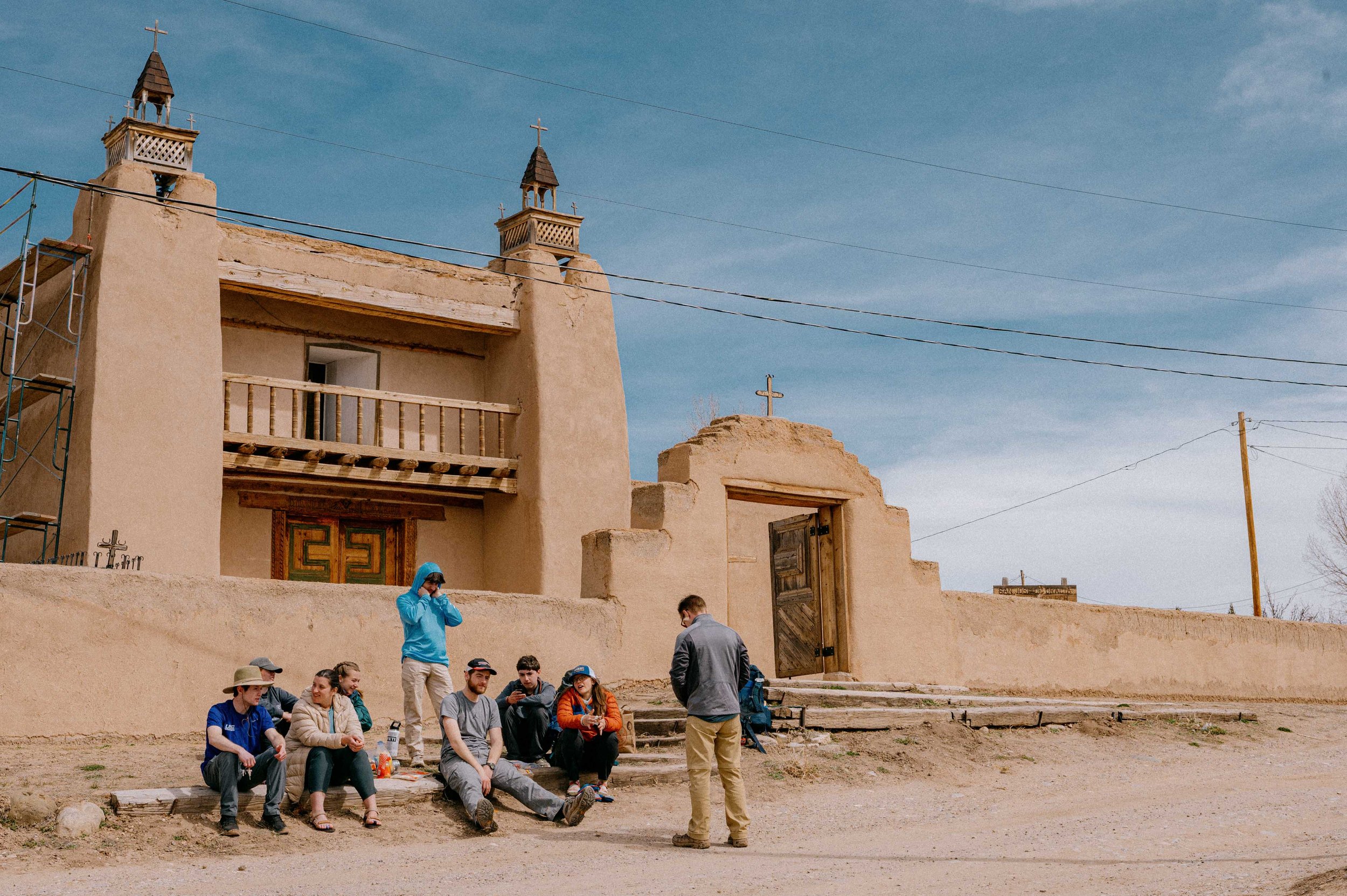 A group of people sitting and standing in front of a building with adobe walls and a wooden door, under a blue sky with power lines. The building features small towers with crosses on top.