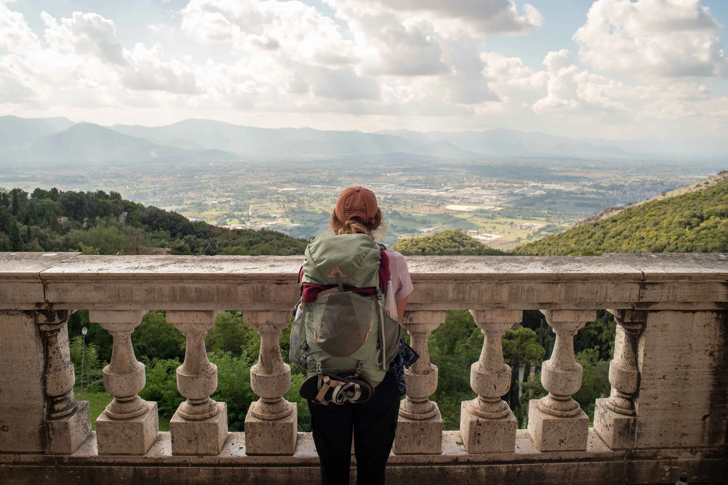 A woman with a large backpack looking out over a scenic landscape from a stone balcony, with mountains and a cloudy sky in the background.
