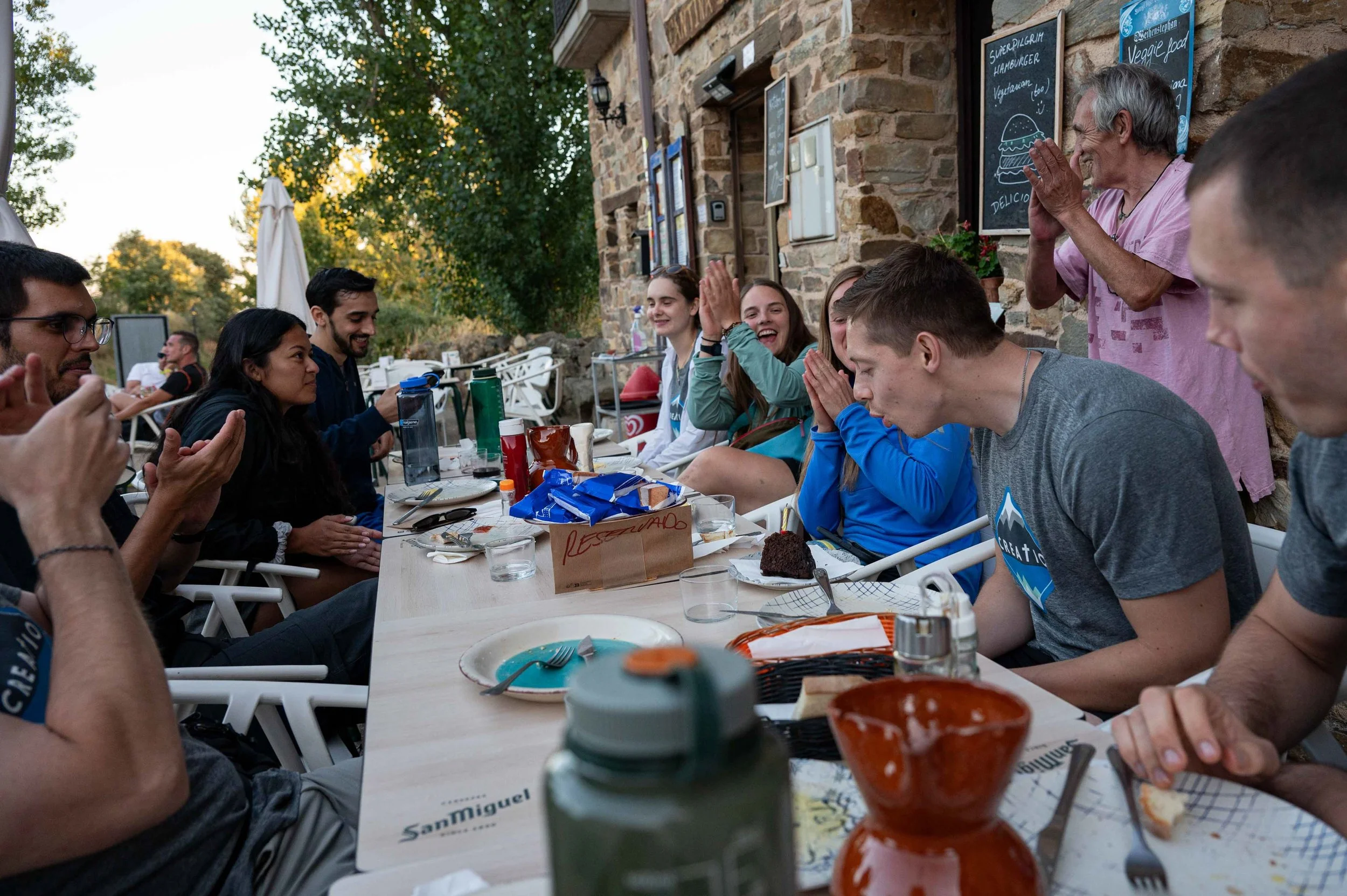 People sitting at an outdoor restaurant table, some smiling and clapping, celebrating an event, with a server standing nearby clapping. The scene appears to be in the evening with trees and a stone building in the background.