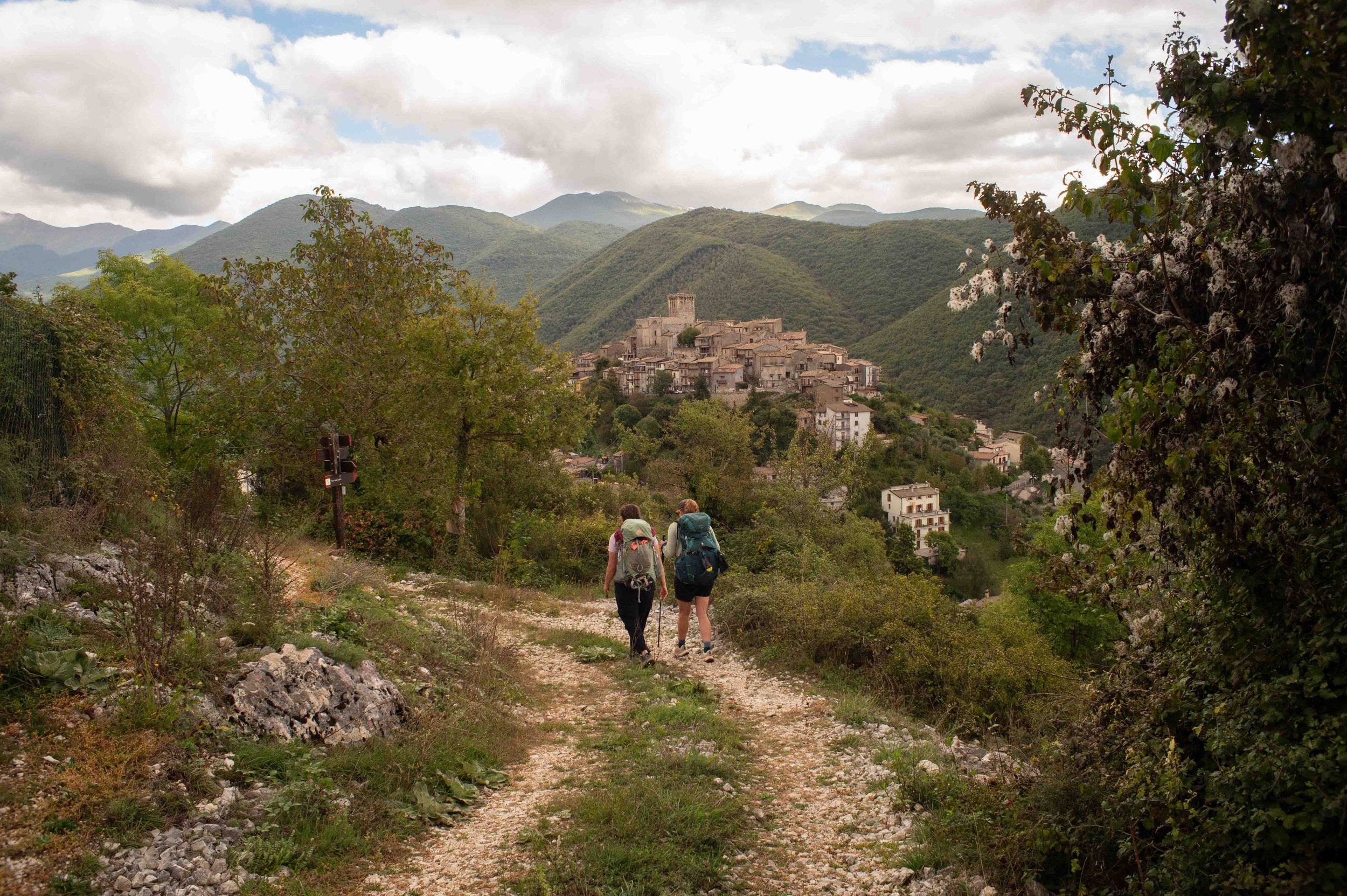 Two hikers with backpacks walk along a dirt trail through greenery with a town on a hill and mountains in the background.